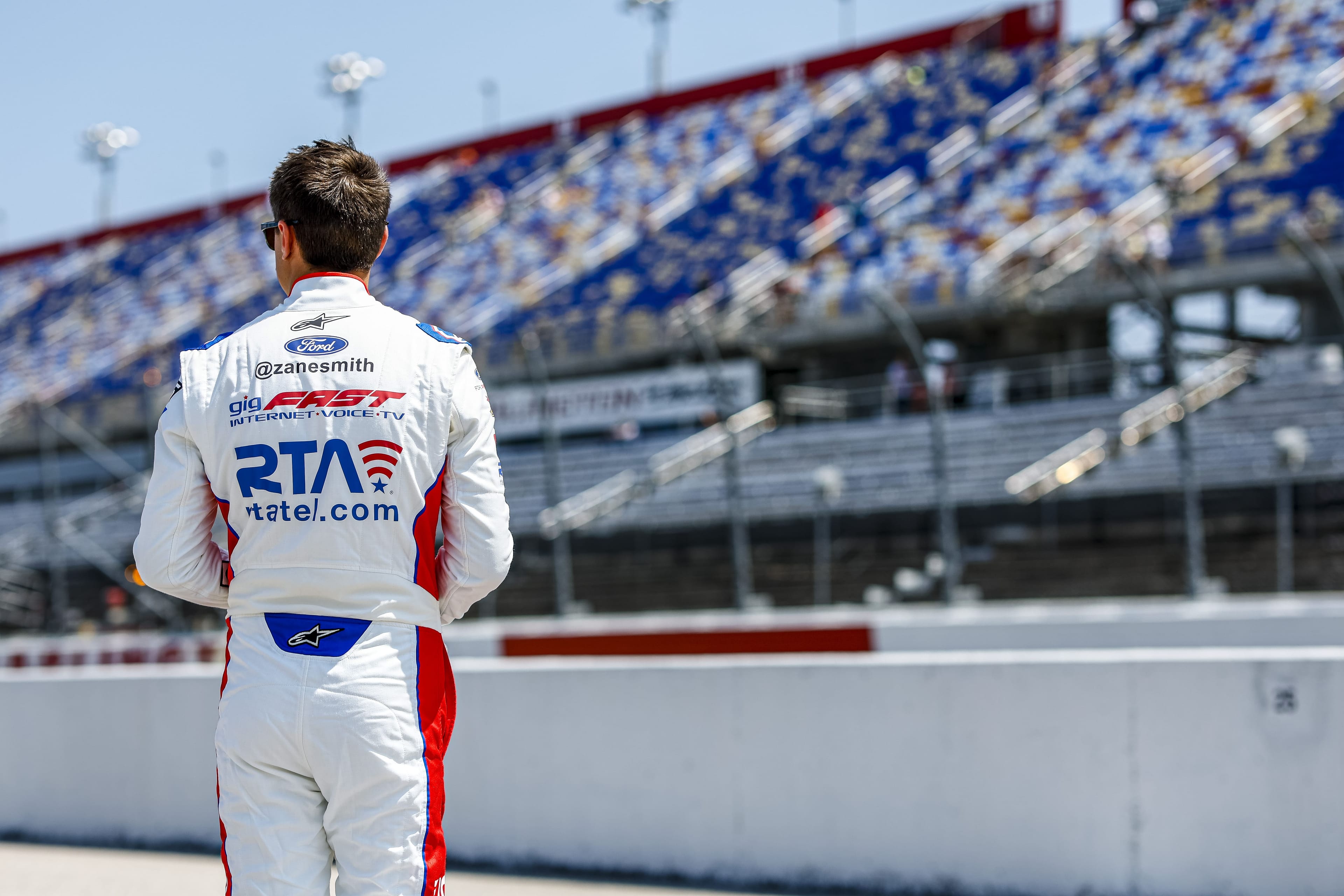 Zane Smith back-facing portrait, racing suit with RTA and gigFAST logos, full stadium backdrop