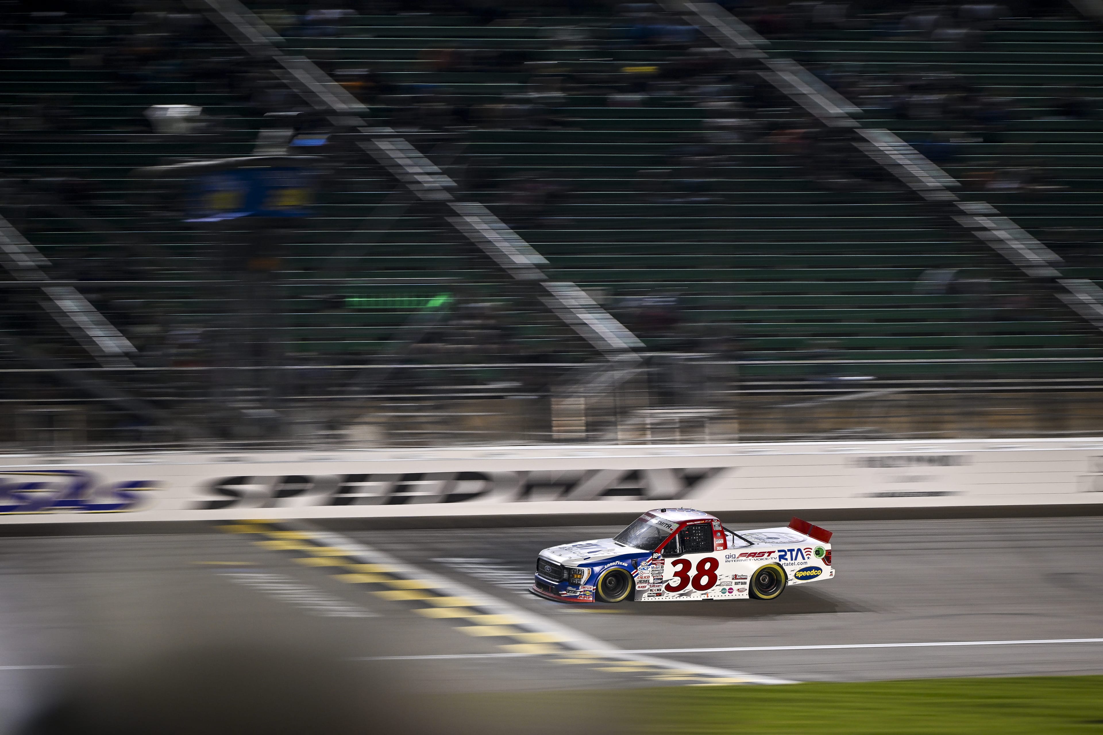 #38 truck at night, passing SPEEDWAY signage, cinematic low-angle shot