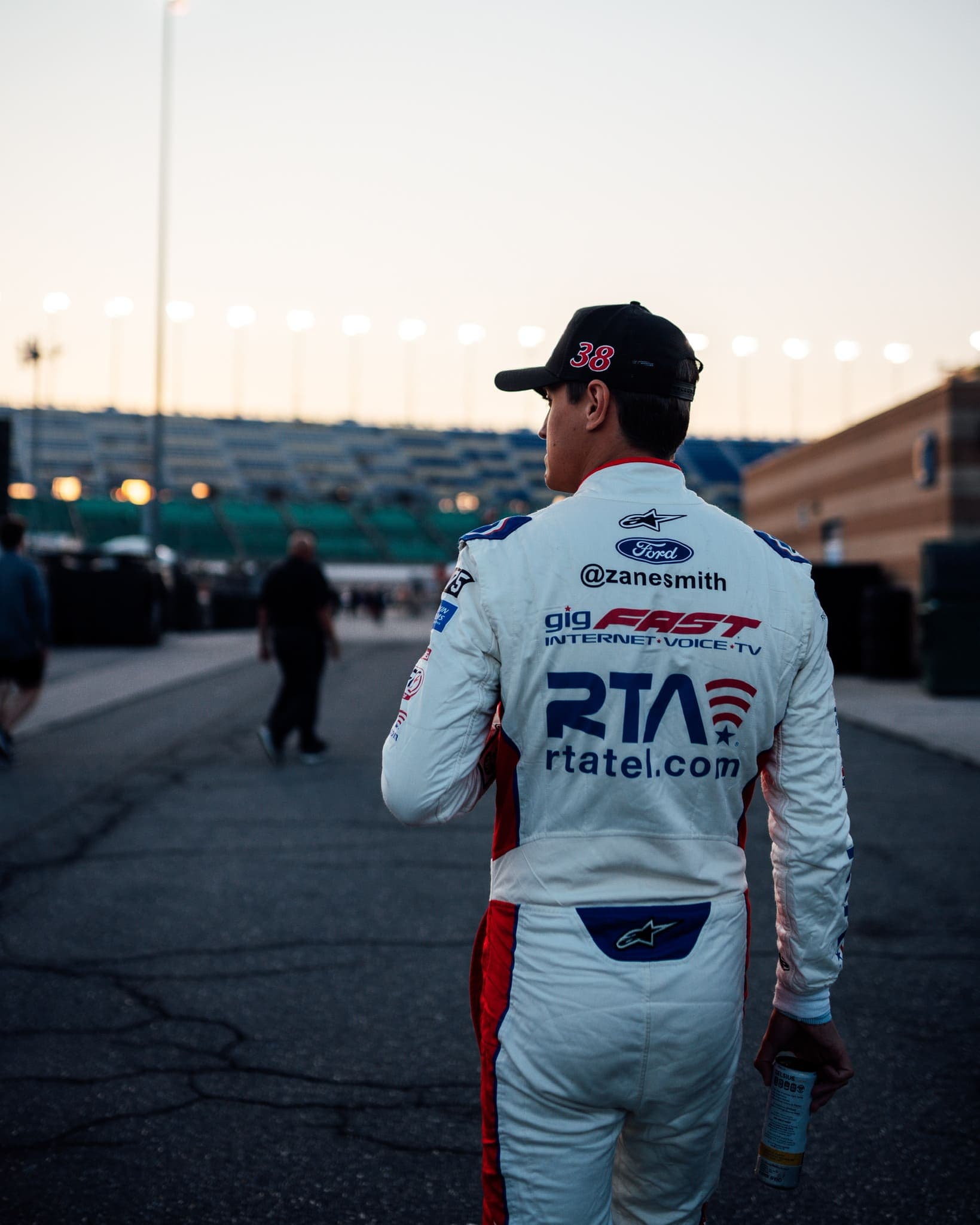 Zane Smith walking at the racetrack at dusk, gigFAST Internet and RTA logos visible on the back of his racing suit